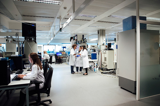 Mixed ethnicity female science research students discuss procedure in a laboratory. University Graduates are mentored by their teacher, they are working on computers and surrounded by state of the art machines for discovering disease. Nuclear magnetic resonance equipment, pioneers disease discovery in fields such as cancer, diabetes, dementia and autism.