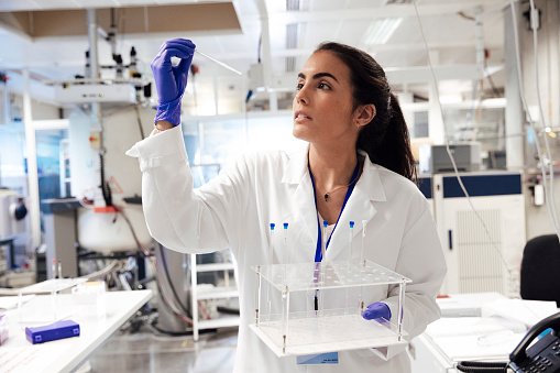 A female scientist examines samples in a laboratory, full of, state of the art research equipment. The NMR apparatus is discovering diseases such as autism, diabetes, dementia and cancer. This biochemist is holding and looking at samples.