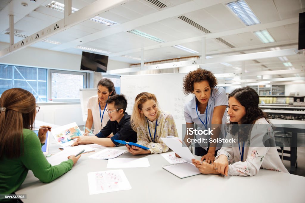 Diverse Females Involved in STEM Teacher with a group of university students, in a laboratory classroom. The instructor is considering one of the students work, the mood is light hearted and positive. Other classmates are discussing things with each other. This is a realistic teaching scenario, with candid expressions. This is a multi-ethnic group of women. In the background there is a white board with mathematical formula written on it. All ladies are wearing id tags. Teacher Stock Photo