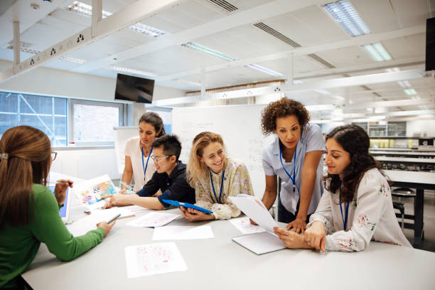 Diverse Females Involved in STEM Teacher with a group of university students, in a laboratory classroom. The instructor is considering one of the students work, the mood is light hearted and positive. Other classmates are discussing things with each other. This is a realistic teaching scenario, with candid expressions. This is a multi-ethnic group of women. In the background there is a white board with mathematical formula written on it. All ladies are wearing id tags. tutor stock pictures, royalty-free photos & images