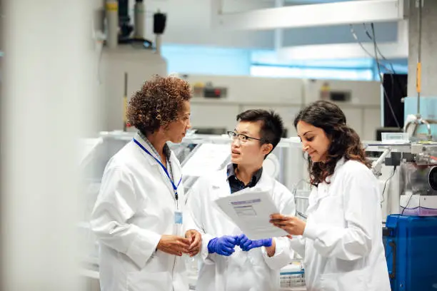 Diverse mix of technicians, wearing lab coats, debate working procedure, in a disease research facility. Nuclear magnates resonance equipment, is in the background, this high end apparatus can be used to discover information on conditions such as dementia, diabetes, autism and cancer. Focus technique and layers, ensure the group of female science graduates and manager are the centre of attention. This is a realistic scenario.