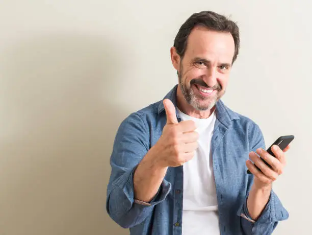 Senior man using smartphone happy with big smile doing ok sign, thumb up with fingers, excellent sign