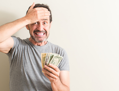 Senior man holding dollar money stressed with hand on head, shocked with shame and surprise face, angry and frustrated. Fear and upset for mistake.