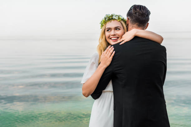 istock attractive smiling bride in wreath and groom in suit hugging on beach 1044249012