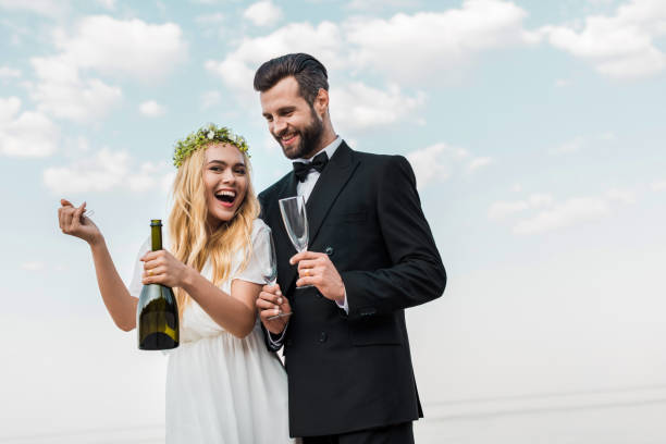 istock laughing bride in white dress opening champagne bottle on beach 1044249042
