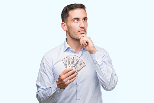 Handsome young man holding money serious face thinking about question, very confused idea