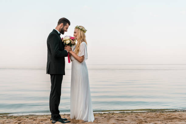 istock side view of affectionate wedding couple standing with bouquet on beach, bride sniffing roses 1044262652