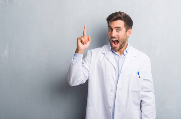 istock Handsome young professional man over grey grunge wall wearing white coat pointing finger up with successful idea. Exited and happy. Number one. 1044295230