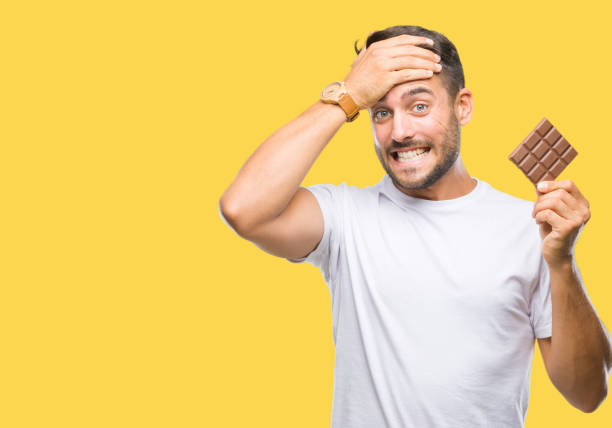 istock Young handsome man eating chocolate bar over isolated background stressed with hand on head, shocked with shame and surprise face, angry and frustrated. Fear and upset for mistake. 1044299152