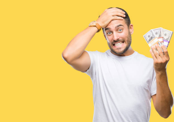 istock Young handsome man holding stack of dollars over isolated background stressed with hand on head, shocked with shame and surprise face, angry and frustrated. Fear and upset for mistake. 1044299296