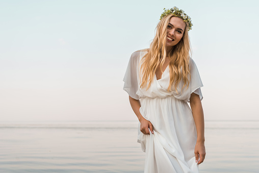 smiling attractive bride in white dress and wreath of flowers looking at camera near ocean