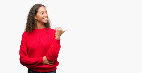 Young hispanic woman wearing red sweater smiling with happy face looking and pointing to the side with thumb up.