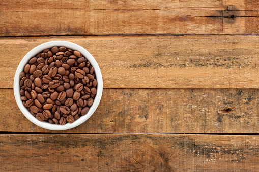 Top view of white bowl full of roasted coffee beans over wooden table