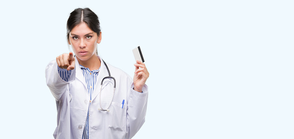 Young hispanic doctor woman holding credit card pointing with finger to the camera and to you, hand sign, positive and confident gesture from the front