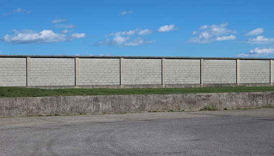 Urban street background. An asphalt road in front of a long concrete brick wall under a blue sky with fluffy clouds