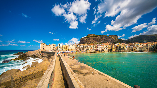 Cefalu, Sicily, Italy - August 13, 2017: view showing Cefalu bridge with people sitting and walking sorrounded by the sea, buildings and rock mountains can be seen on the background