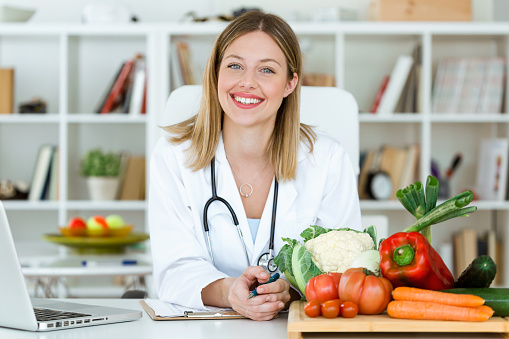 Hermosa nutricionista sonriente mirando a cámara y mostrando verduras saludables en la consulta. photo