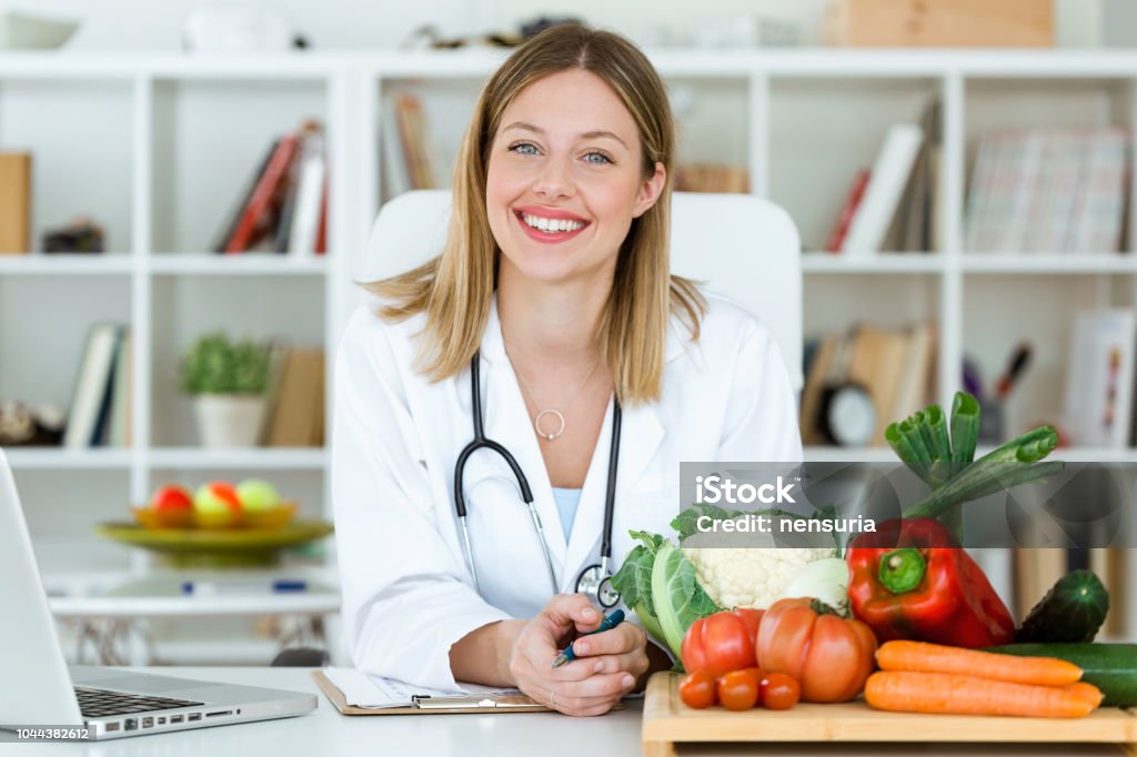 Bellissimo nutrizionista sorridente che guarda la macchina fotografica e mostra verdure sane nella consultazione. - Foto stock royalty-free di Nutrizionista