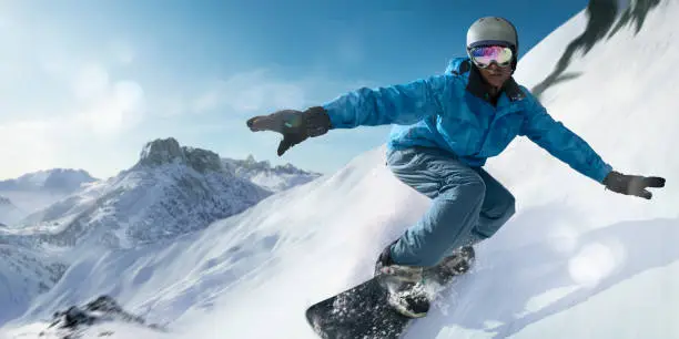 A close up image of a black snowboarder laying into a carve turning the snowboard towards the camera. The snowboarder is dressed in blue jacket, salopettes, helmet and visor and is travelling at high speed down a slope with snow spraying out from behind the board. With motion blur.