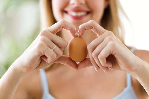 Shot of beautiful young woman showing brown chicken egg with hands in a heart shape at home.