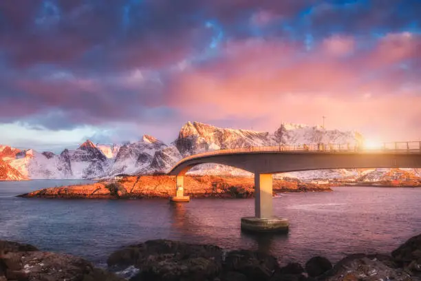 Beautiful bridge and snowy mountains at sunrise in winter in Lofoten islands, Norway. Landscape with bridge, colorful sky with pink clouds, rocks in snow, stones in the sea and sun. Travel in europe