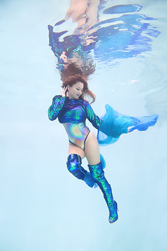 An underwater portrait of a woman wearing green and blue sequined stilletto boots and matching shirt.