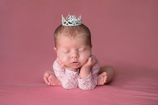 A beautiful, sleeping, newborn baby girl wearing a rhinestone tiara. Shot in the studio with a pink backdrop.
