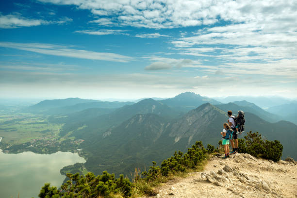 Walchensee, Germany - August 23, 2017: Man with daughter and a baby over his back stands on Herzogstand mountain top looking at a view featuring mountain ridge, valley and a lake