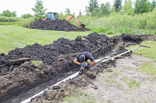 Erskine, USA - June 25, 2018:  A new residential mound septic system is being installed. A worker is installing the septic pipe in the trench while another worker in the background is  operating a backhoe to prepare the ground for the drain field.
