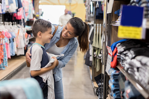 Happy young boy looks at a shirt in the mirror while shopping for back to school clothes with his mom.