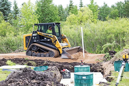 Erskine, USA - June 25, 2018:  A new residential mound septic system is being installed. Scene of the construction site.  A worker is operating a bobcat to apply a bed of sand on the drain field.