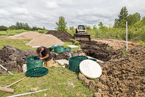 Erskine, USA - June 25, 2018:  A new residential mound septic system is being installed. Scene of a construction site where a new septic tank has been lowered into the ground.  A worker is installing the risers - covers.