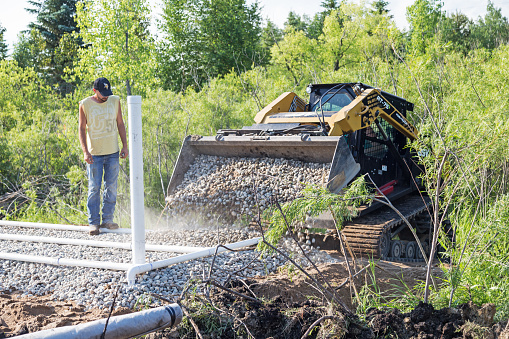 Erskine, USA - June 26, 2018: A new residential mound septic system is being installed. A barely visible worker operating a bobcat is applying rock on top of drain field pipes as another worker looks on.