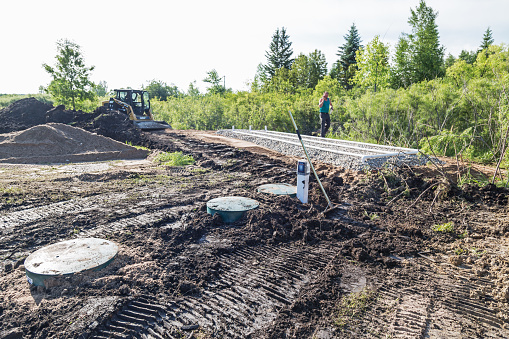 Erskine, USA - June 26, 2018: A new residential mound septic system is being installed. Construction site scene showing dirt covering the newly installed septic tank, a PS Patrol System Monitoring alarm, and the drain field.  In the background, a worker is walking while talking on his mobile phone.