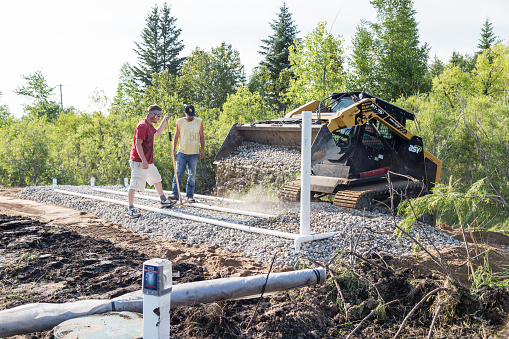 Erskine, USA - June 26, 2018: A new residential mound septic system is being installed. A barely visible worker operating a bobcat is applying rock on top of drain field pipes while two other workers look on.