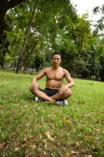 młody dorosły azjatycki mężczyzna ćwiczący i relaksujący się w publicznym parku - zen like nature breathing exercise sitting zdjęcia i obrazy z banku zdjęć