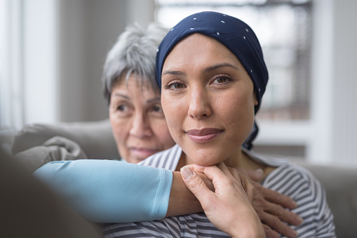 An ethnic woman wearing a headscarf and fighting cancer sits on the couch with her mother. She is in the foreground and her mom is behind her, with her arm wrapped around in an embrace. She is looking at the camera with an expression of resolute confidence and serenity.