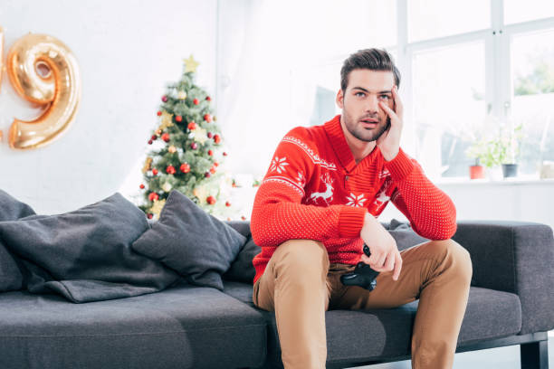 istock upset man holding joystick and sitting on sofa with christmas tree behind 1044646024