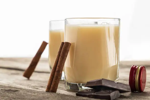 Milk iqueur in two glasses with cinnamon and chocolate, on wooden table. White background. Copy space