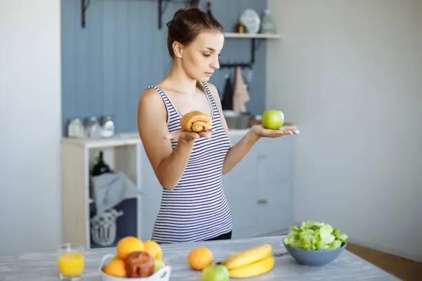 Photo of Woman holding in hand cake sweet bun and apple fruit choosing, trying to resist temptation, making the right dietary choice.
