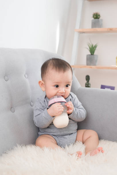 piccola ragazza carina seduta in camera sul divano a bere latte dalla bottiglia e sorridente. bambino felice. concetti interni per famiglie. l'infanzia è il momento migliore! - sucking asian ethnicity baby mother foto e immagini stock