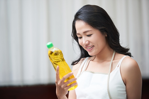 Beautiful Asian woman in white apron holding bottle of yellow detergent and reading label