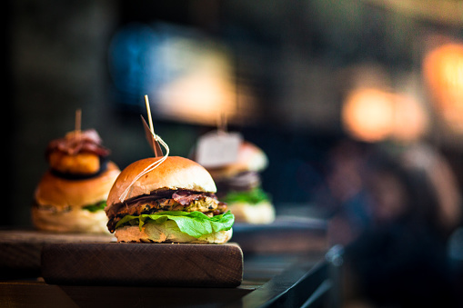 Close up image of a selection of freshly flame grilled burgers in a row on a wooden counter at a London street food market. Each of the burgers has its own label, on which is written the contents of the burger. The burgers are sandwiched between glazed buns, and presented on beds of fresh green lettuce and stuffed with melted cheese and red onion. Horizontal colour image with copy space and beautiful bokeh background.
