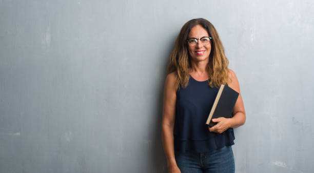 middle age hispanic woman standing over grey grunge wall holding a book with a happy face standing and smiling with a confident smile showing teeth - women professor mature adult human face imagens e fotografias de stock