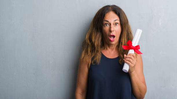 istock Middle age hispanic woman standing over grey grunge wall holding diploma scared in shock with a surprise face, afraid and excited with fear expression 1044762064