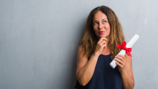 istock Middle age hispanic woman standing over grey grunge wall holding diploma serious face thinking about question, very confused idea 1044762164