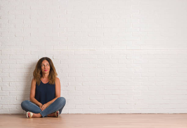 istock Middle age hispanic woman sitting on the floor over white brick wall smiling looking side and staring away thinking. 1044763352