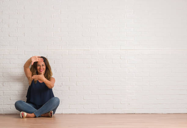 istock Middle age hispanic woman sitting on the floor over white brick wall smiling making frame with hands and fingers with happy face. Creativity and photography concept. 1044763534