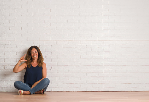 Middle age hispanic woman sitting on the floor over white brick wall smiling and confident gesturing with hand doing size sign with fingers while looking and the camera. Measure concept.