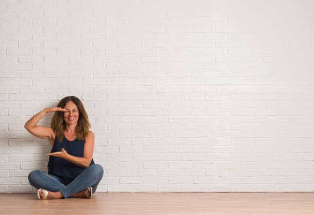 istock Middle age hispanic woman sitting on the floor over white brick wall gesturing with hands showing big and large size sign, measure symbol. Smiling looking at the camera. Measuring concept. 1044763616
